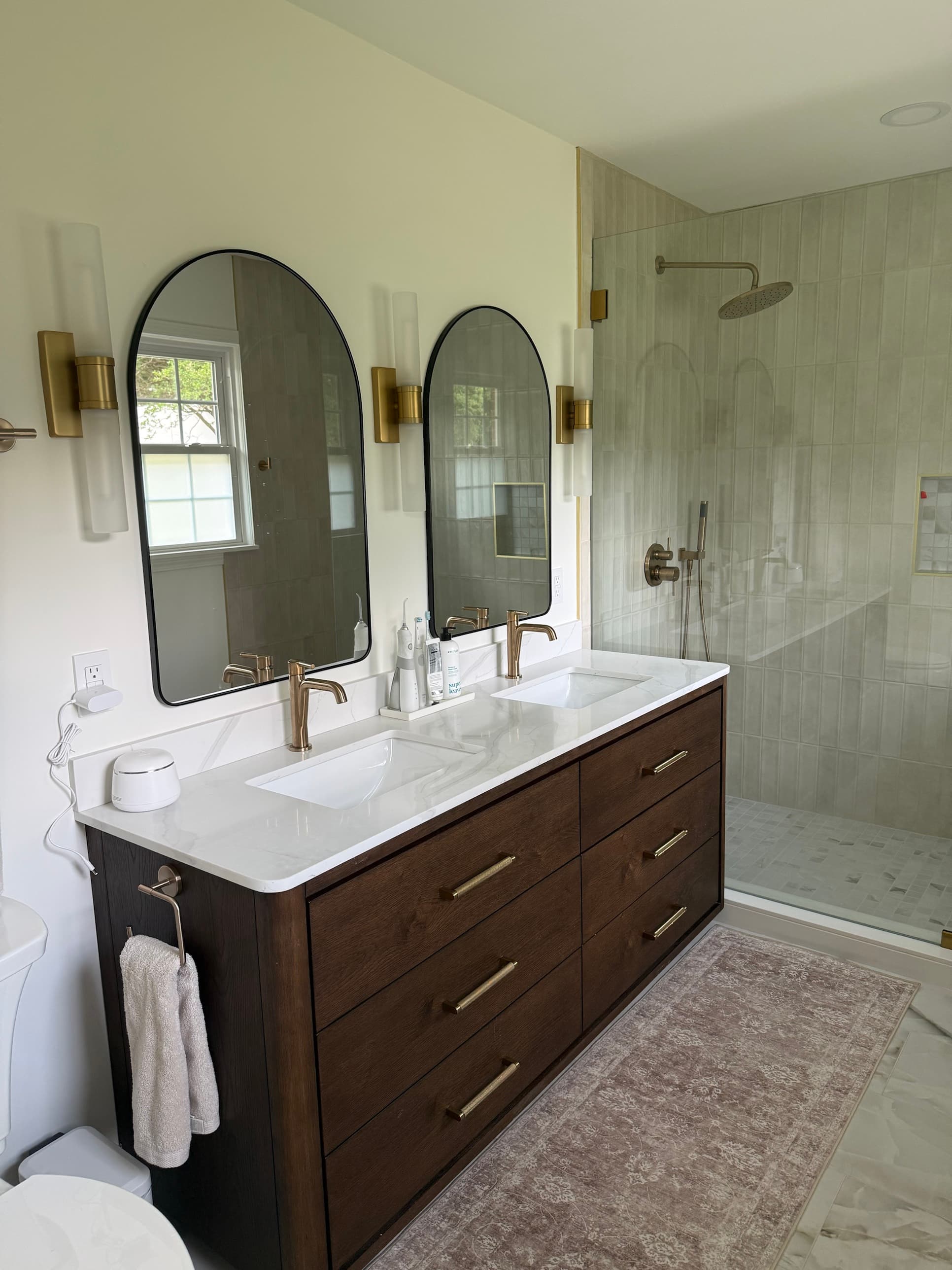 Primary bathroom renovation with dark wood double vanity, arched mirrors, gold sconces, and walk-in tile shower by Remodel CLT in Charlotte, NC